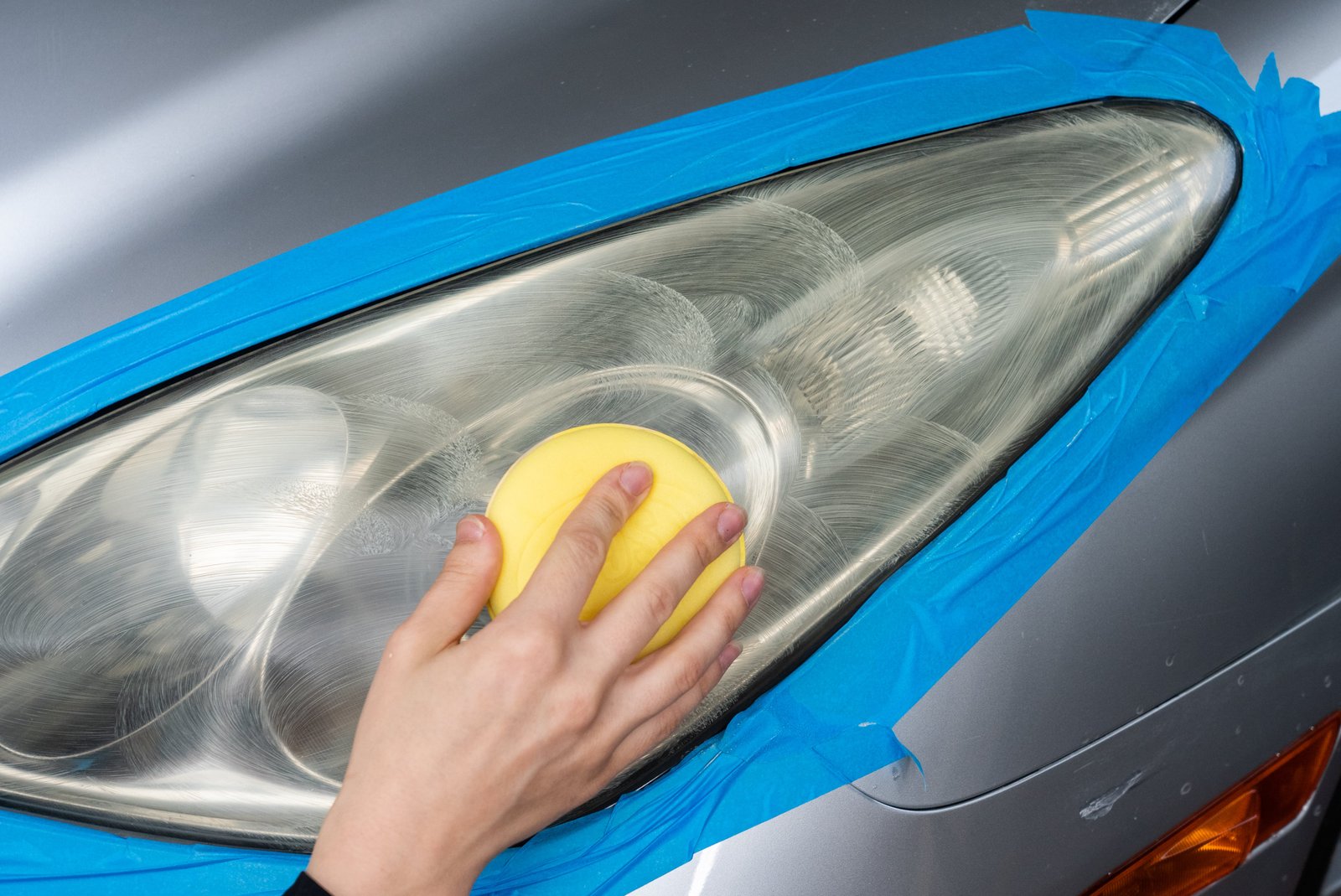 Close-up of a cloudy headlight being restored with a polishing pad, edges carefully masked with blue painter's tape